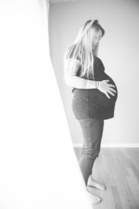 Black and white photo of a standing pregnant woman standing near a window holding her belly.