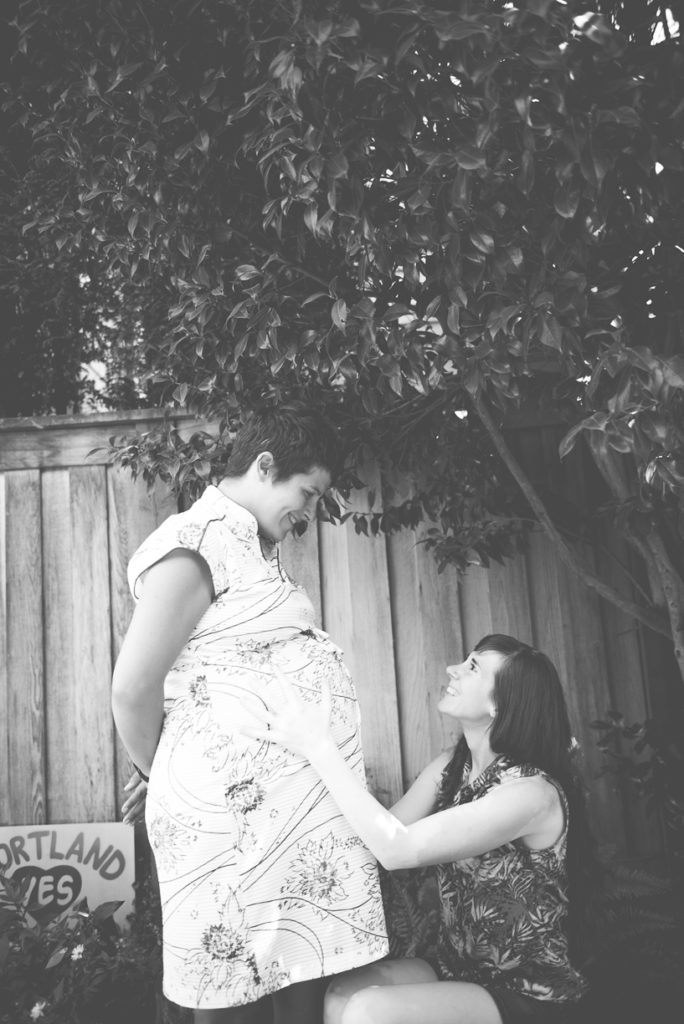 Black and white photo of a standing pregnant woman and her wife sitting and holding her belly.