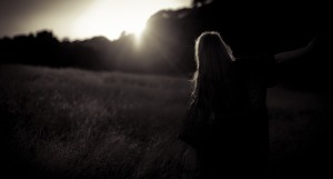 Dark photo of the back of a woman in a field of tall grass looking at the setting sun behind trees.