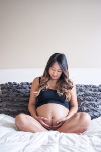 Pregnant woman sitting on her bed looking down at her pregnant belly smiling.