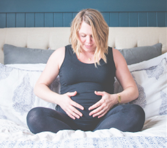 Pregnant woman sitting on her bed holding and looking at her pregnant belly smiling.