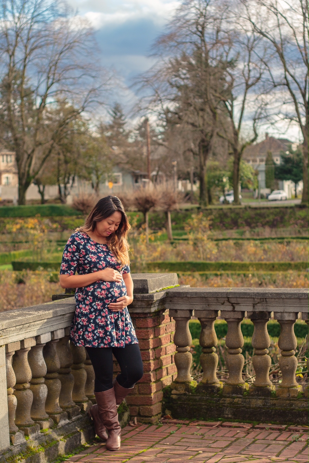 Pregnant woman holding and looking at her pregnant belly, wearing a flower-covered dress, in the rose garden in North Portland, Oregon.